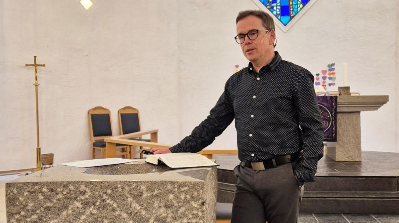 En mann står ved døpefonten i Sund kyrkje og snakker, han har en bok og et papir liggende på døpefonten. Han er kledd i mørk skjorte og grå bukse.