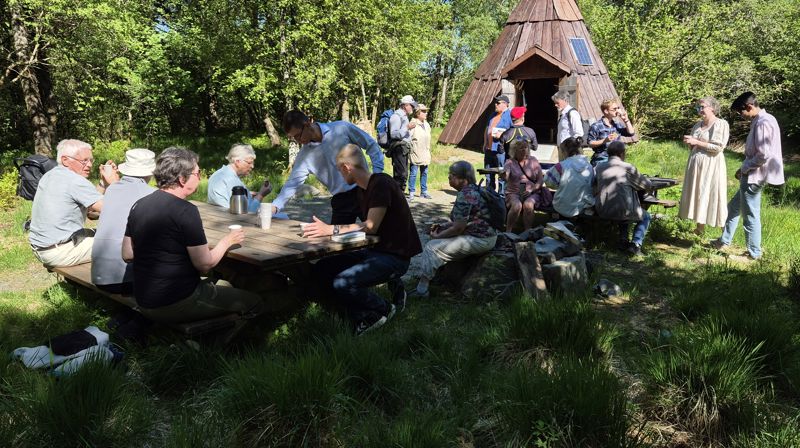 Ute i naturen sitter og står 18 mennesker rundt piknikkbord, i bakgrunnen står en lavvo laget av tre. Det er grønt og sommerlig i naturen og solen skinner