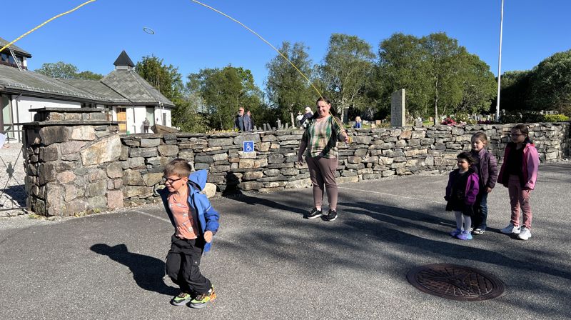 Ute på en plass framfor kirken hoopper en gutt hoppetau, en dame står bak og svinger tauet. til høyre står tre barn og venter på tur. Det er fint vær,.
