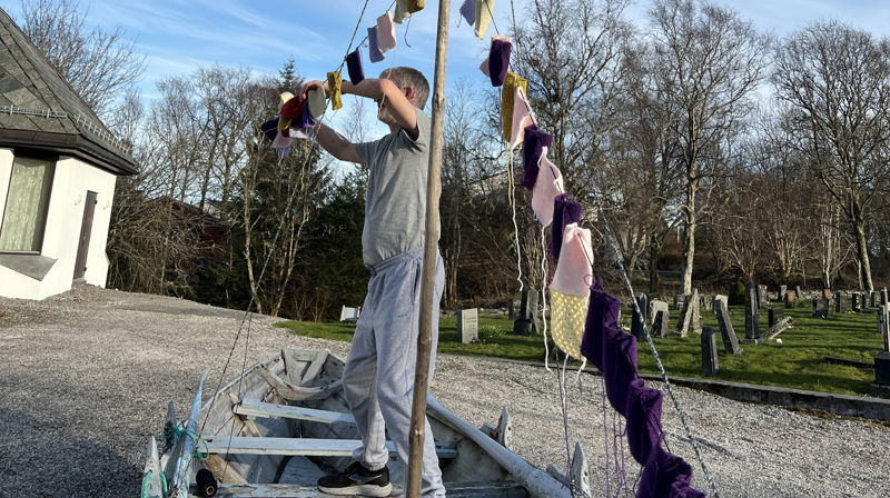 En gammel trebåt stå ute på en grusplass, en mann står oppi båten og henger opp strikke-lapper som er festet som flagg langs et tau. Det er fint ute.