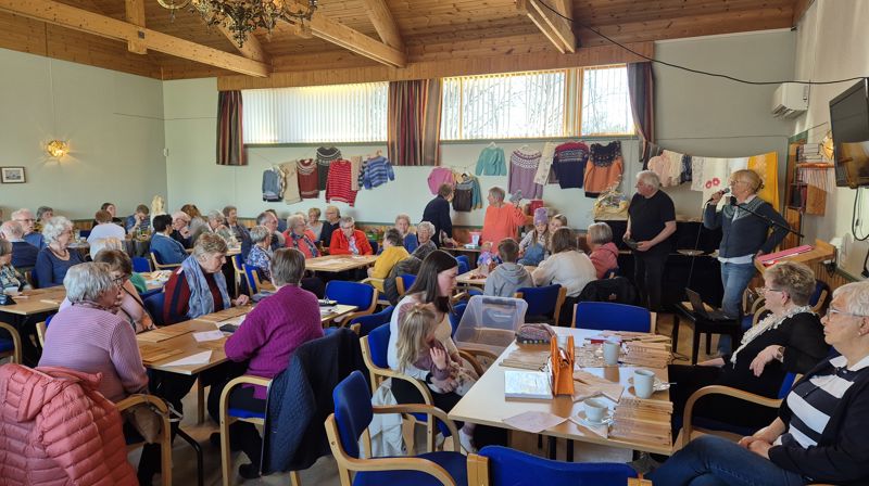 Inne i kyrkjelydshuset er det fullt av mennesker rundt bord, på veggen hhenger mstrikkede gensere, det er basar