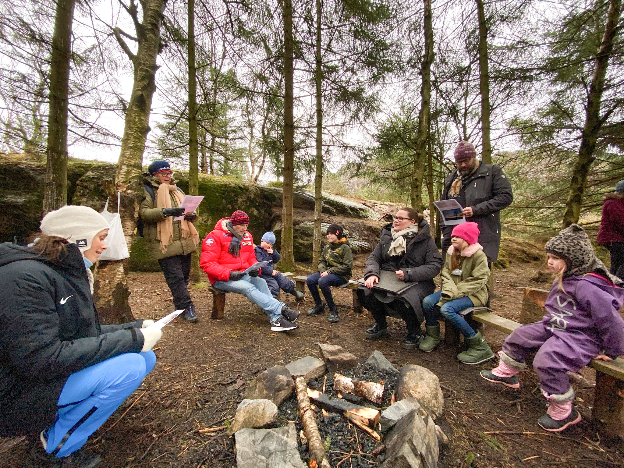 Barn og voksne sitter ute på tømmerbenker rundt et bål, de synger en sang