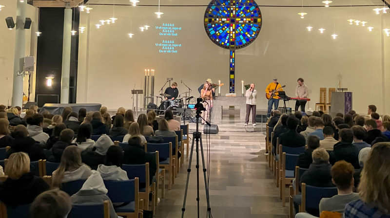 Sett bakfra og frem i kirken, stolene er fulle av ungdommer, fremme står noen og spiller instrumenter og synger