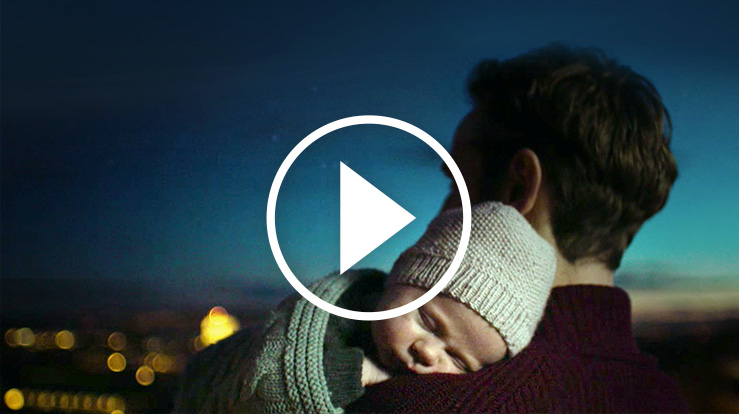 En baby b&#230;res tett inntil brystet av en mann. Det er et &quot;play&quot; symbol foran bilde.